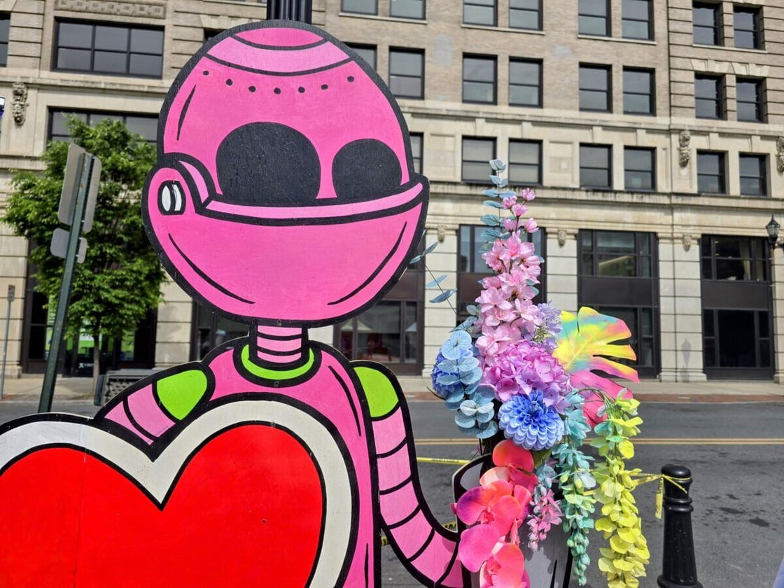 A vibrant pink robot cutout holding a heart, adorned with colorful flowers, stands in front of urban buildings on a sunny day.