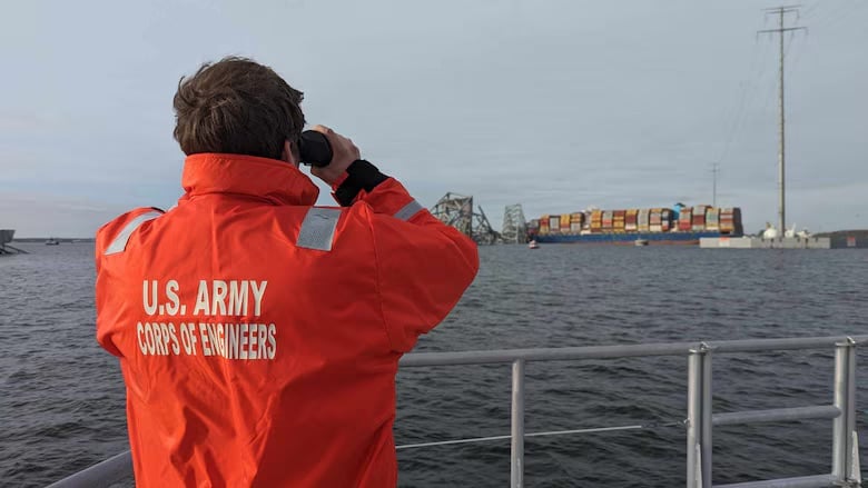 Person in orange jacket looks across the Patapsco River at a multicolored cargo ship near the wreckage of a brown bridge