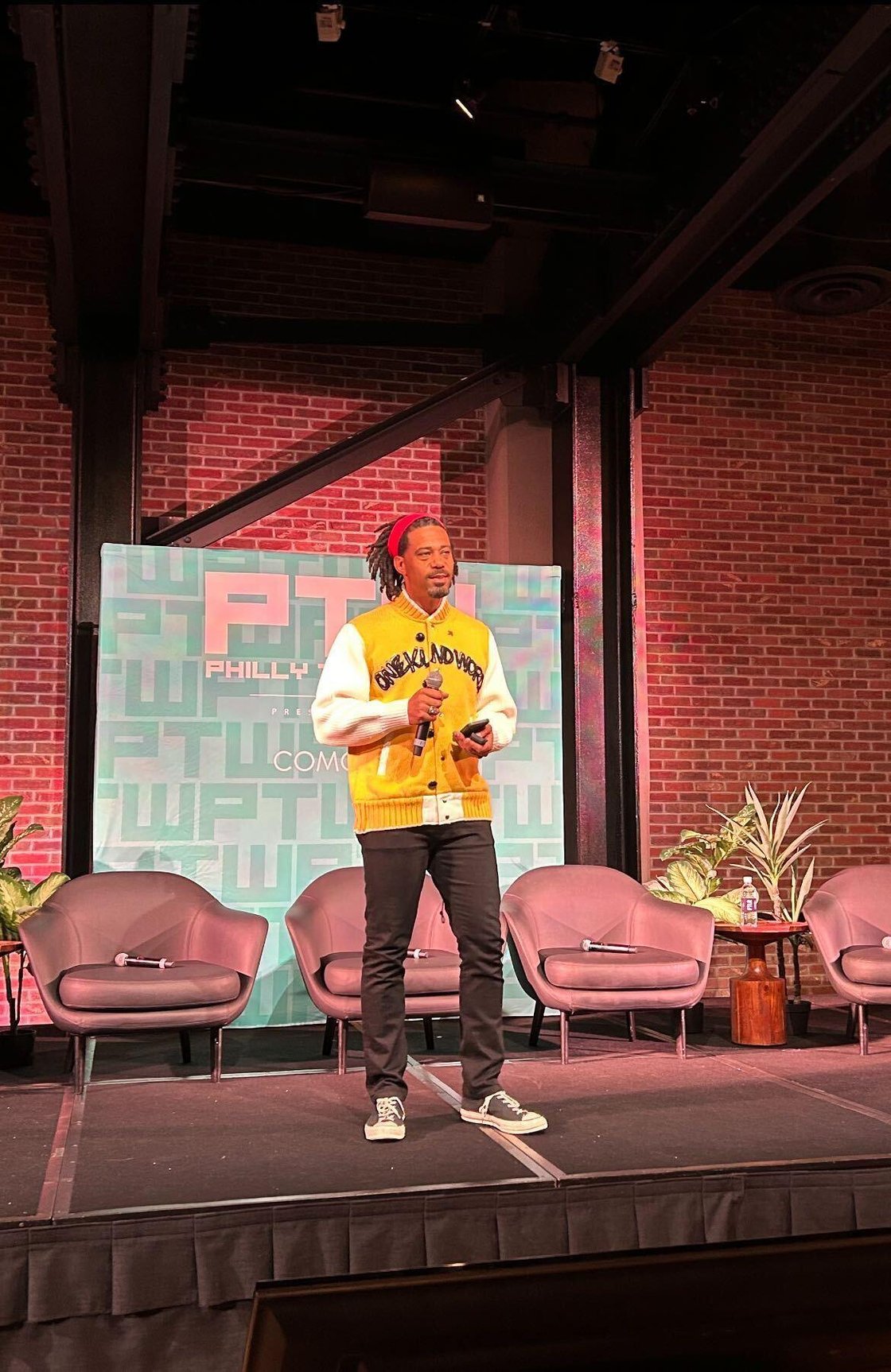 Man in yellow and white jacket and black pants speaks in front of red brick wall.