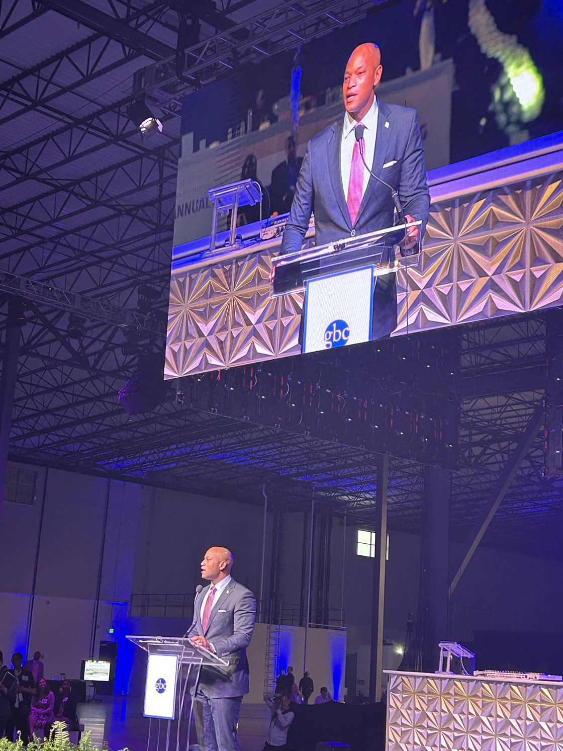 Gov. Wes Moore speaks at a glass podium, with speaking projected on screen hanging from black architecture 