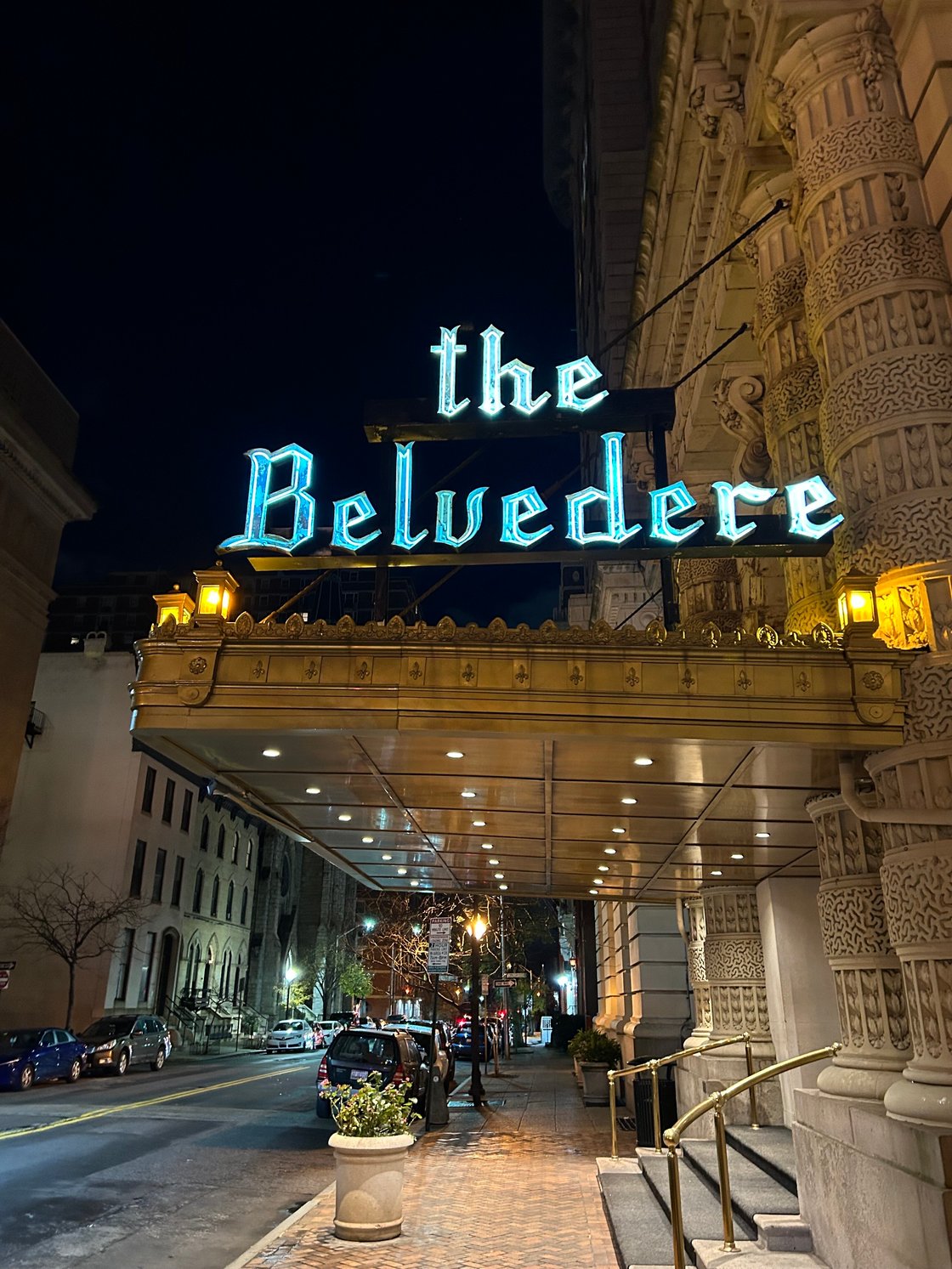 A brown hotel facade and awning bears a blue-lit sign on the top, reading "the Belvedere"