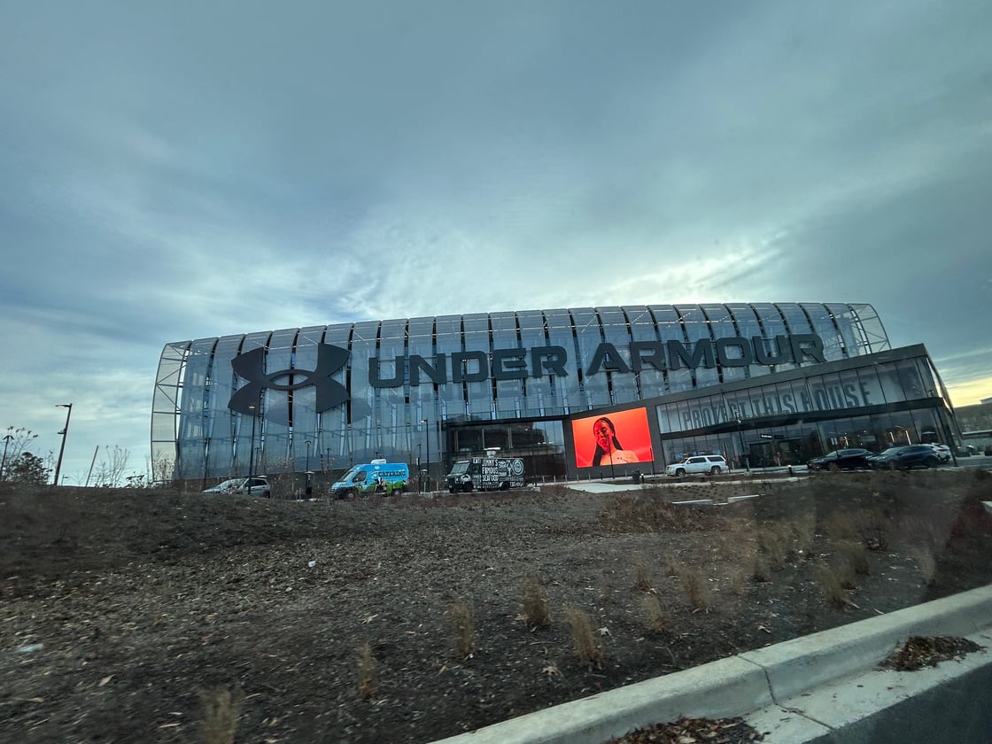 A glass bears a letters reading "UNDER ARMOUR" next to the Under Armour logo and "PROTECT THIS HOUSE". An orange screen depicts a person with long hair in front of the building, with a grey sky in the back.