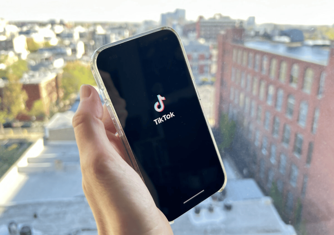 Hand holding a smartphone displaying the TikTok app logo, with a cityscape visible in the blurred background.