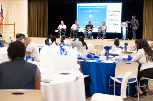 A panel of five people speaks on stage at a tech-focused employer event, with attendees seated at round tables and a presentation screen in the background.