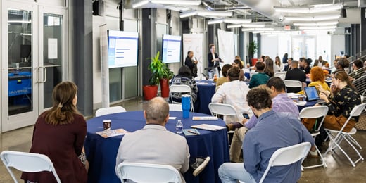 People seated at round tables listen to a presentation in a modern conference room; slides are displayed on large screens at the front.