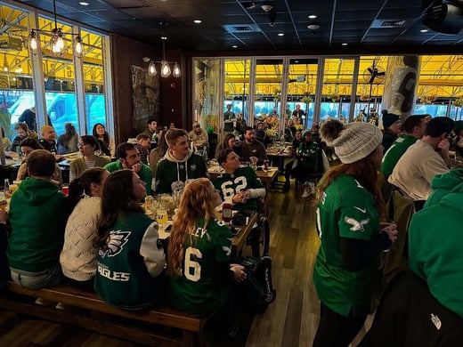 A group of people wearing green football jerseys sit and stand in a restaurant, watching something together, with drinks and food on the tables.