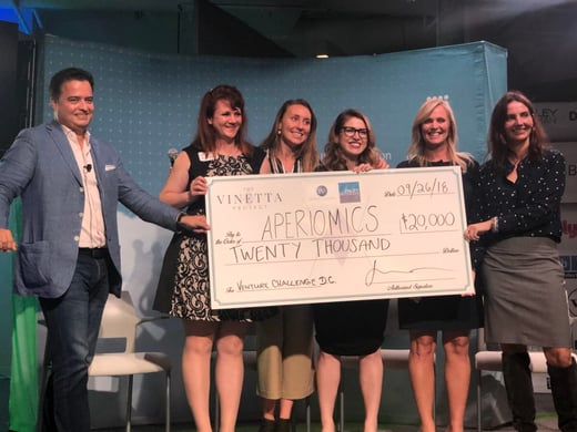 A group of women stands on a stage holding an oversized check awarding them funding for their startup.