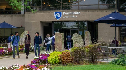 Students walking near the entrance of the Penn State Great Valley campus with the main building and Penn State signage in the background, surrounded by colorful flowers and outdoor seating.