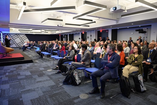 Audience seated in a modern conference room listens to a speaker on stage; some people have backpacks, and cameras are set up in the back.