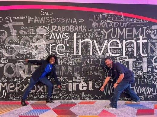 Two people pose in front of a large, chalk-covered AWS re:Invent wall with various signatures and doodles.