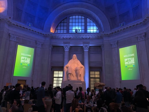Large crowd gathered in a grand hall with a statue of a seated Benjamin Franklin; two green screens display “Welcome PTW 2025.” Blue and purple lights illuminate the space.