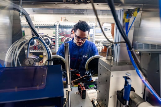 A person in a lab coat and safety glasses works with equipment inside a laboratory glove box surrounded by scientific instruments and wires.