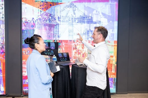 Two people are conversing in front of a vibrant digital display. A man gestures towards the screen, while a woman holding a notepad listens. Laptops are on a table near them.