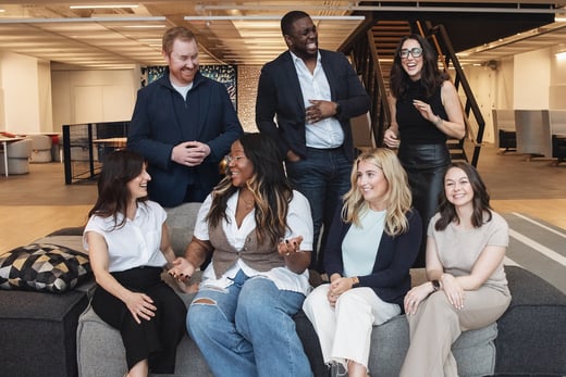Seven adults, four seated and three standing, gather in a modern office space, smiling and interacting with each other in a casual, friendly atmosphere.
