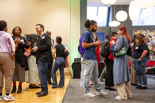 A group of people stand in small clusters, talking and holding drinks at an indoor networking event or social gathering.