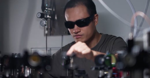 A person wearing dark safety glasses adjusts equipment in a laboratory setting, surrounded by optical instruments and devices.