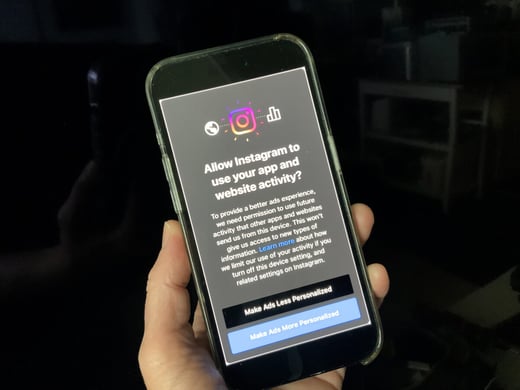 A hand holds a smartphone displaying an Instagram permission screen asking to allow the use of app and website activity for personalized ads, with options to make ads less or more personalized.