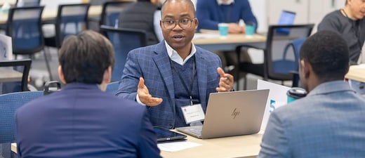A man in a suit speaks to two other men across a table in an office setting, with laptops and papers visible.