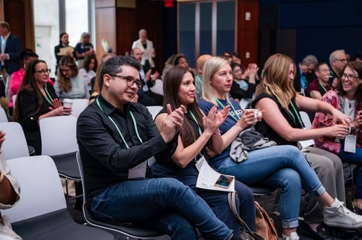Audience members seated indoors clap and smile during a conference or seminar, with name badges and programs visible.