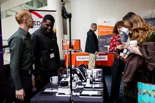 A group of people chat near a demonstration of black and white devices and an orange table.