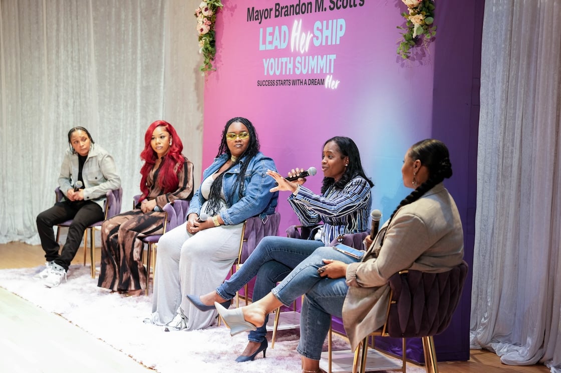 Five Black women sit on chairs in front of a pink and blue screen and white curtain during panel