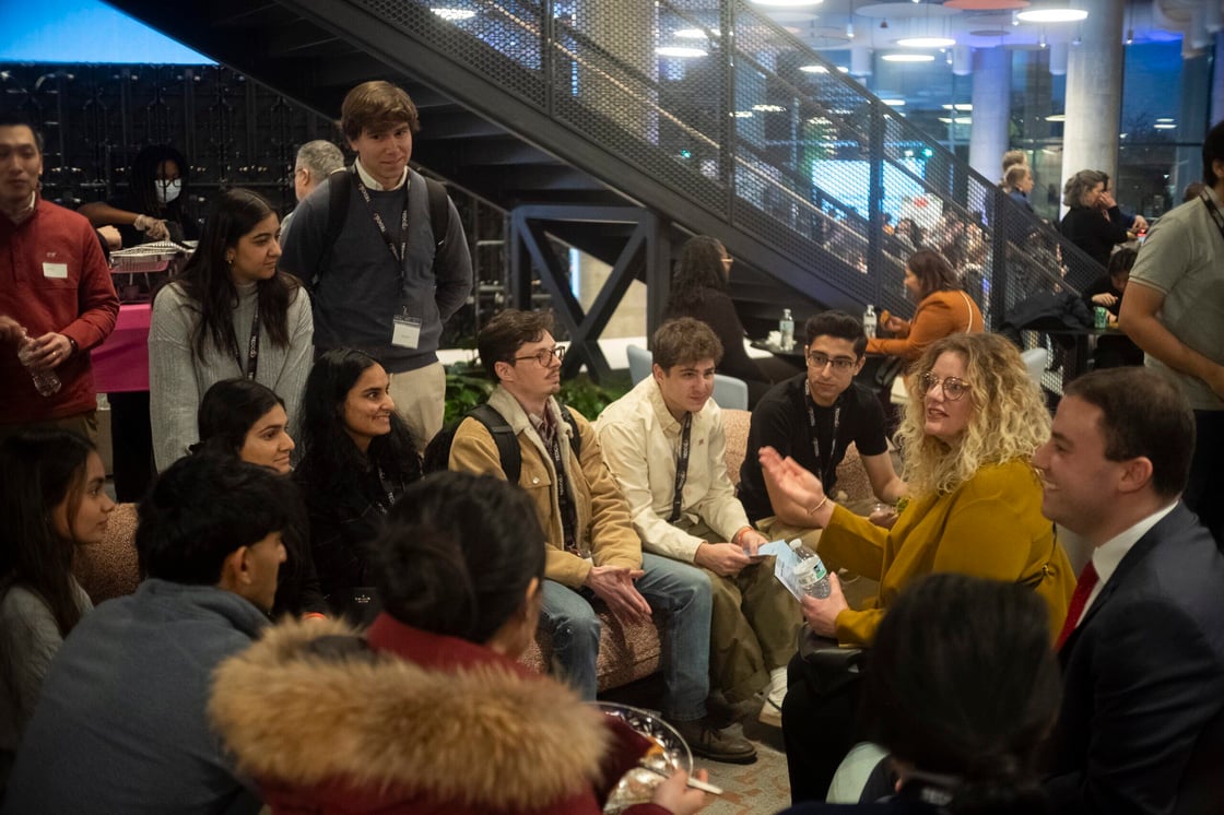A woman with blonde hair and glasses speaks to a group of seated and standing young people in front of a dark grey metal stairwell and glass windows.
