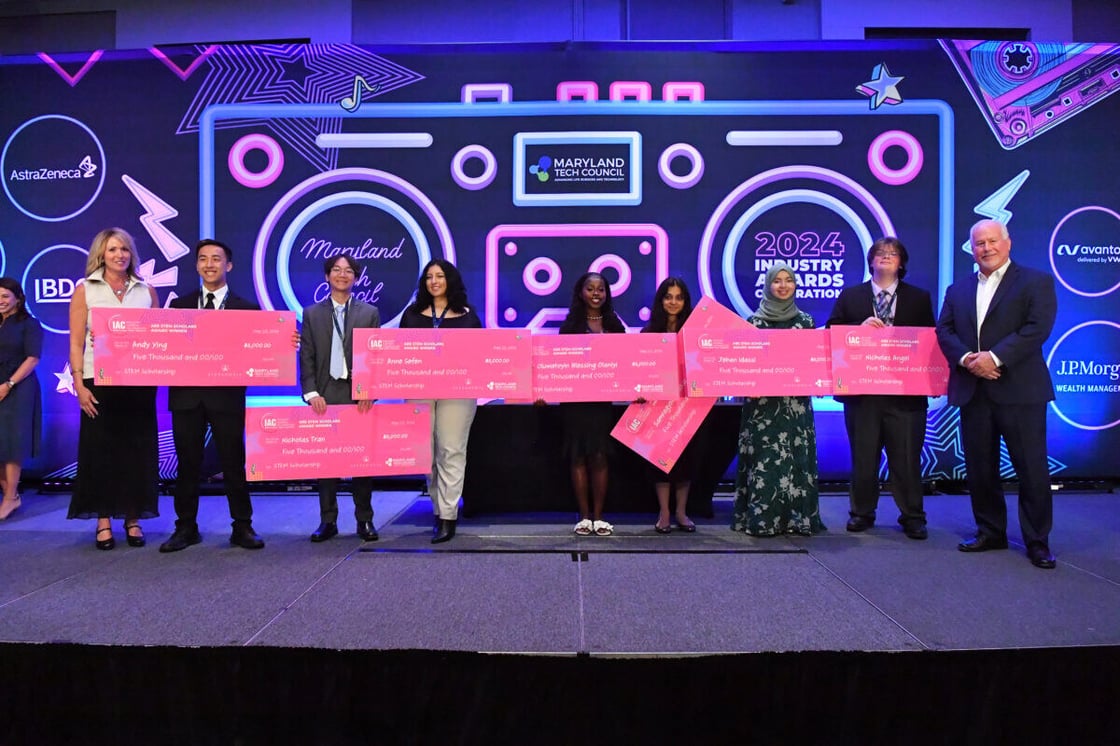 Seven young people hold pink novelty checks while flanked by two adults in front of neon stereo.