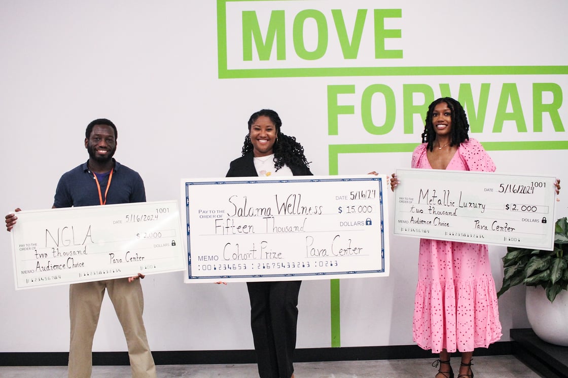 A Black man and two Black women hold oversized checks in front of a white wall that reads 