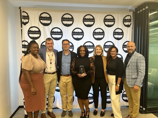 A group of seven people standing together in front of a logo backdrop, smiling and posing for a photo. One person in the center is holding an award.