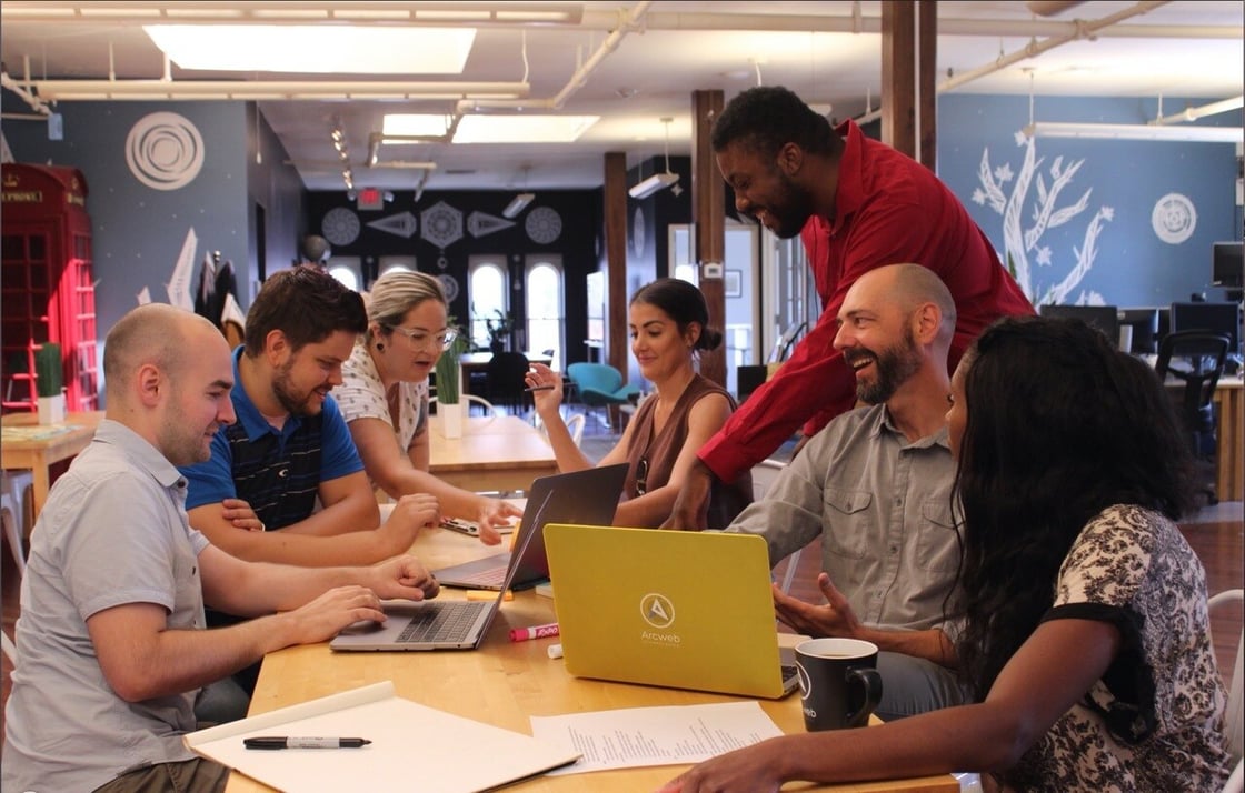 Six seated people work around laptops while another stands