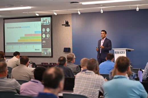 A presenter speaks to an audience in a conference room, standing by a podium and a screen displaying a technology evolution chart.