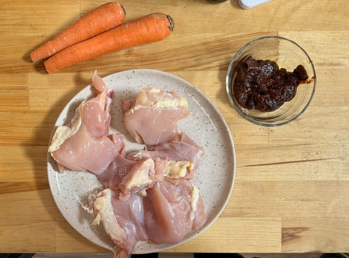 Plate of raw chicken next to two whole carrots and a small bowl of a dark brown sauce on a wooden surface.