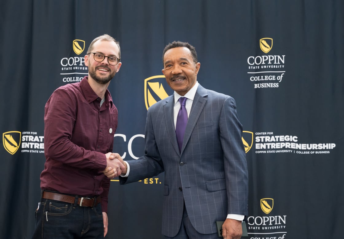 Men shake hands in front of black curtain with white and yellow Coppin State branding