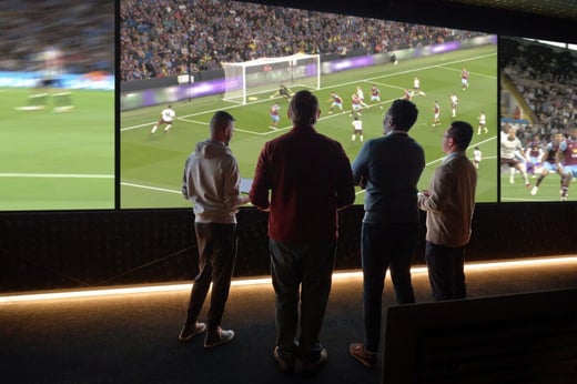 Four people stand inside, watching a soccer match on a large screen displaying blurred players and a goal in progress.