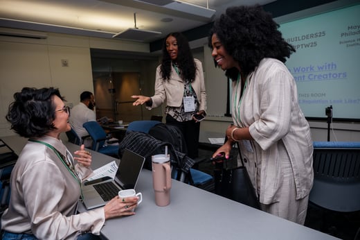 Three women converse and smile in a conference room, with laptops, coffee cups, and event badges visible on a table.